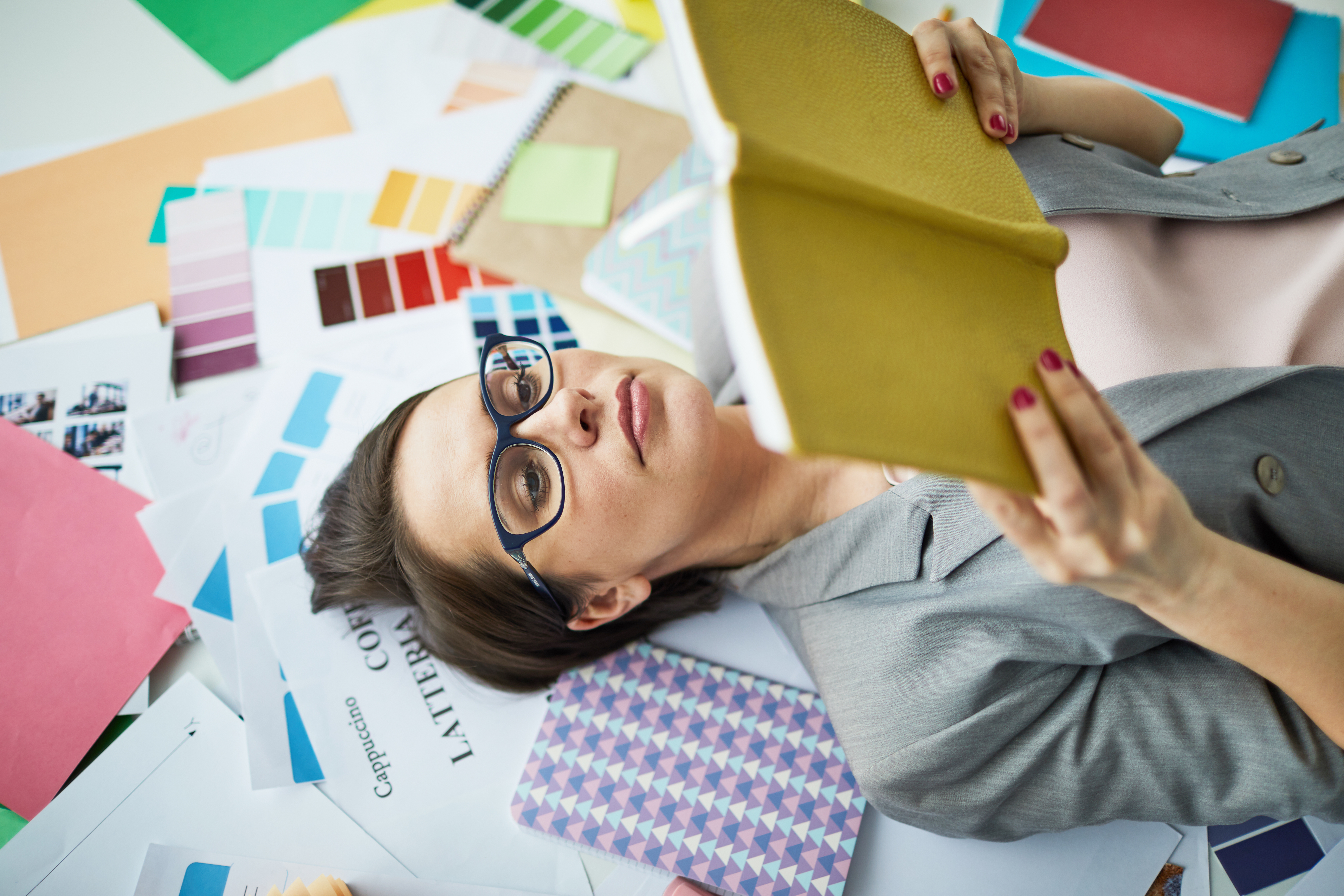 Top view portrait of contemporary businesswoman wearing glasses  reading book while  lying over  art and design supplies, copy space