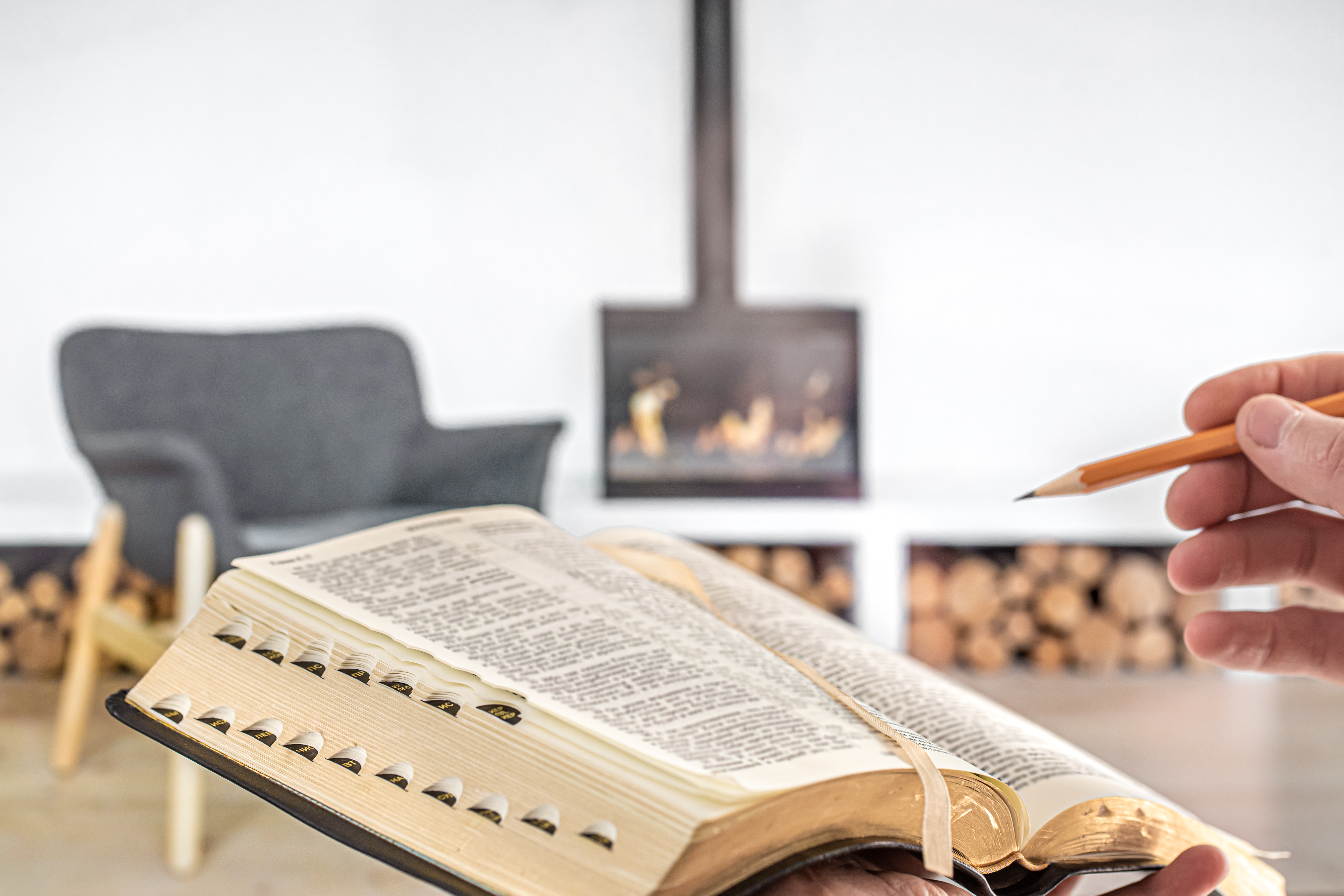 A man holding a Bible with a pencil, against the background of the living room with a fireplace. Reading a book in a cozy environment. Close up.